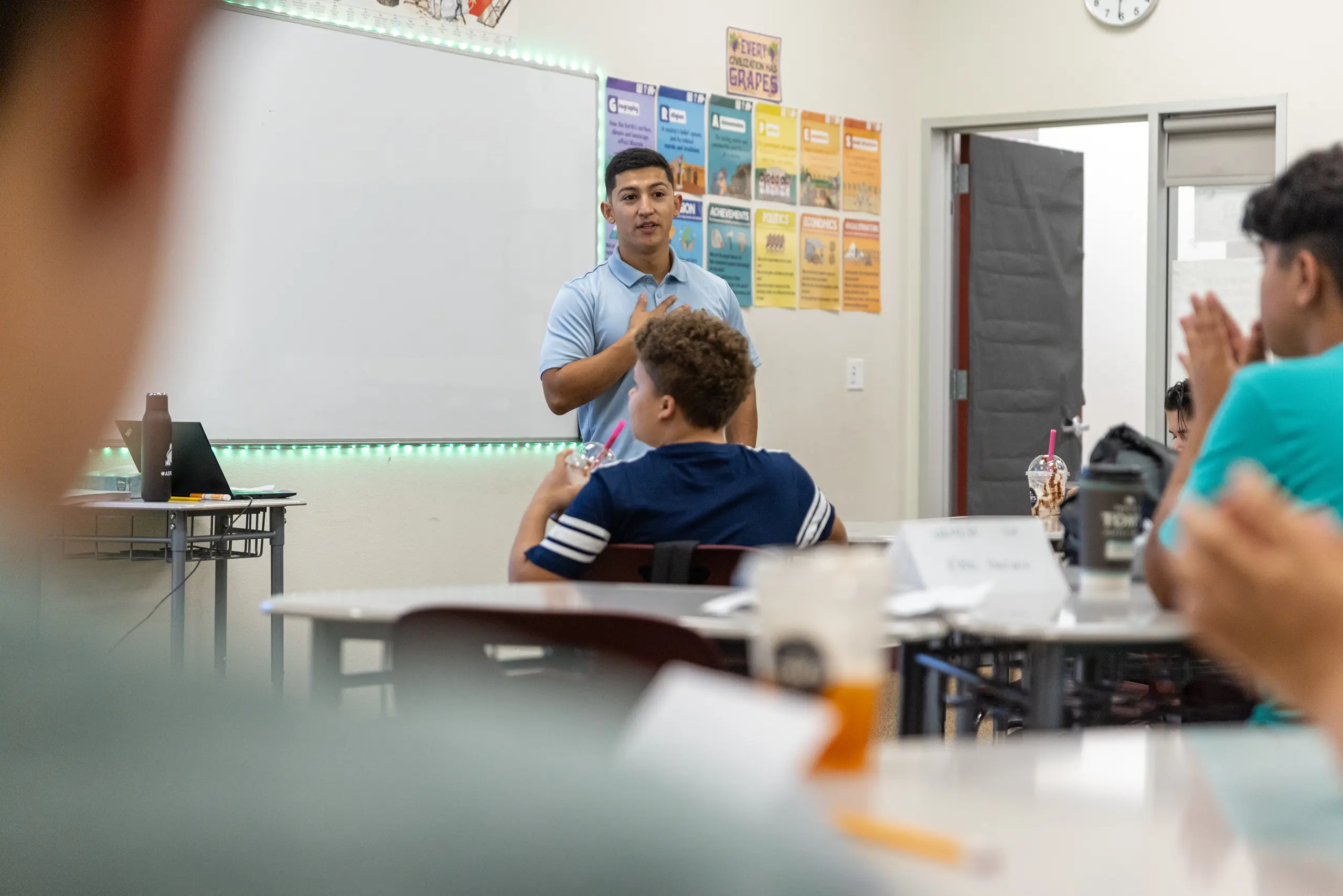 A teacher stands at the front of a classroom speaking to students, who are seated at desks. Colorful posters are on the wall behind him, and a whiteboard is visible. The students are listening attentively.