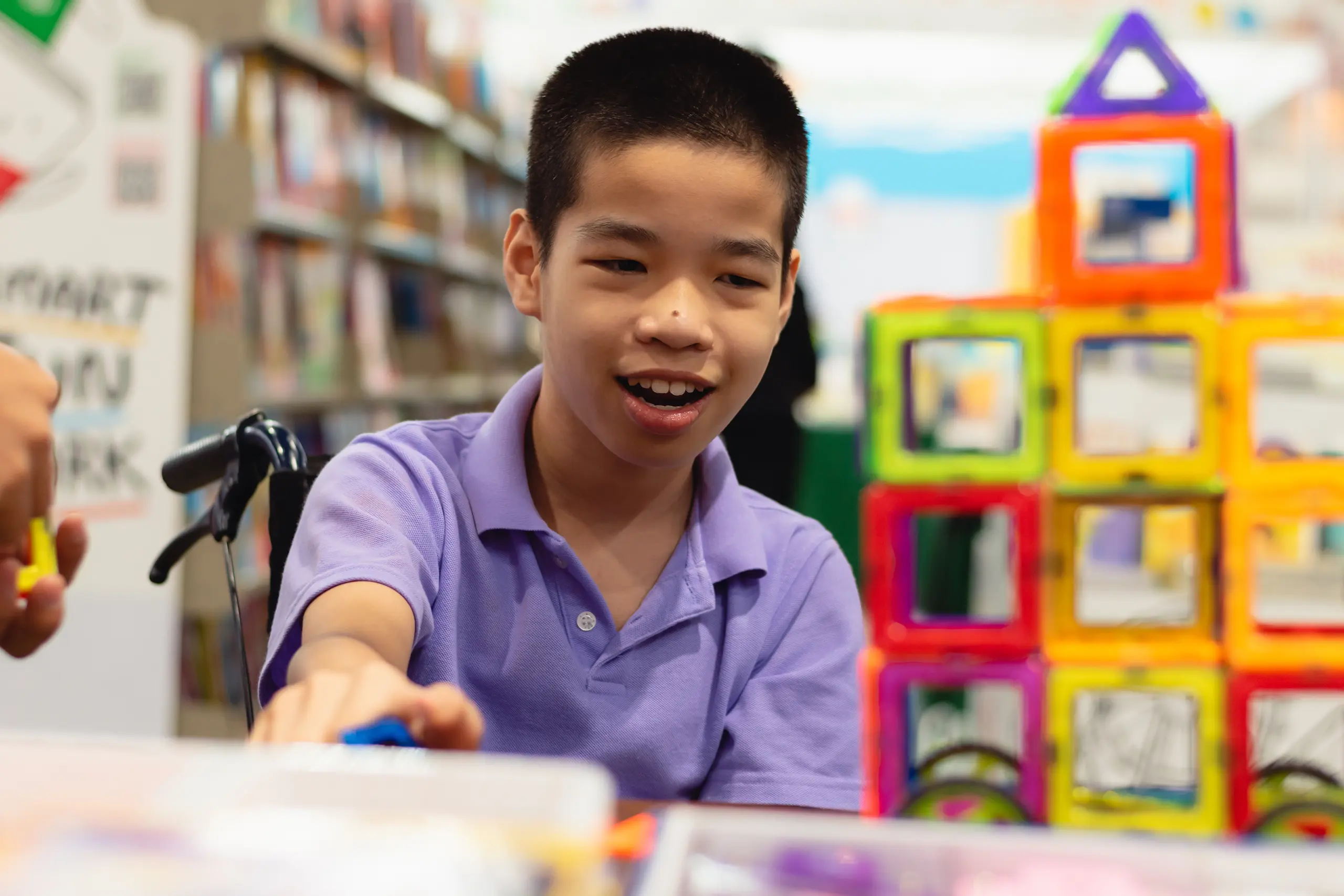 A smiling boy wearing a purple shirt sits in a library, reaching toward colorful magnetic building blocks stacked into a structure on the table in front of him. Shelves of books are visible in the background.