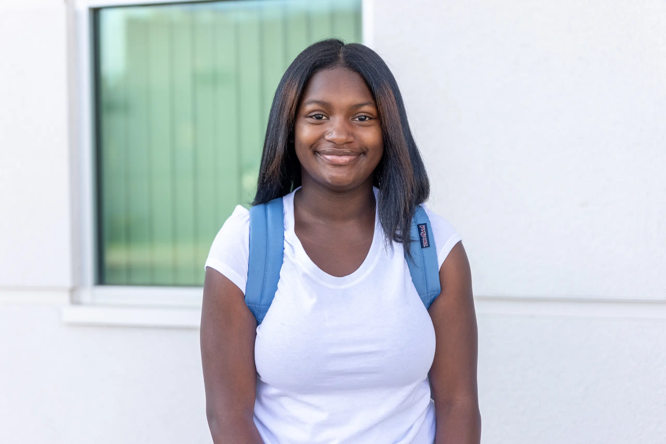 A young woman wearing a white t-shirt and blue backpack stands in front of a window, smiling at the camera. The background is a light-colored building.