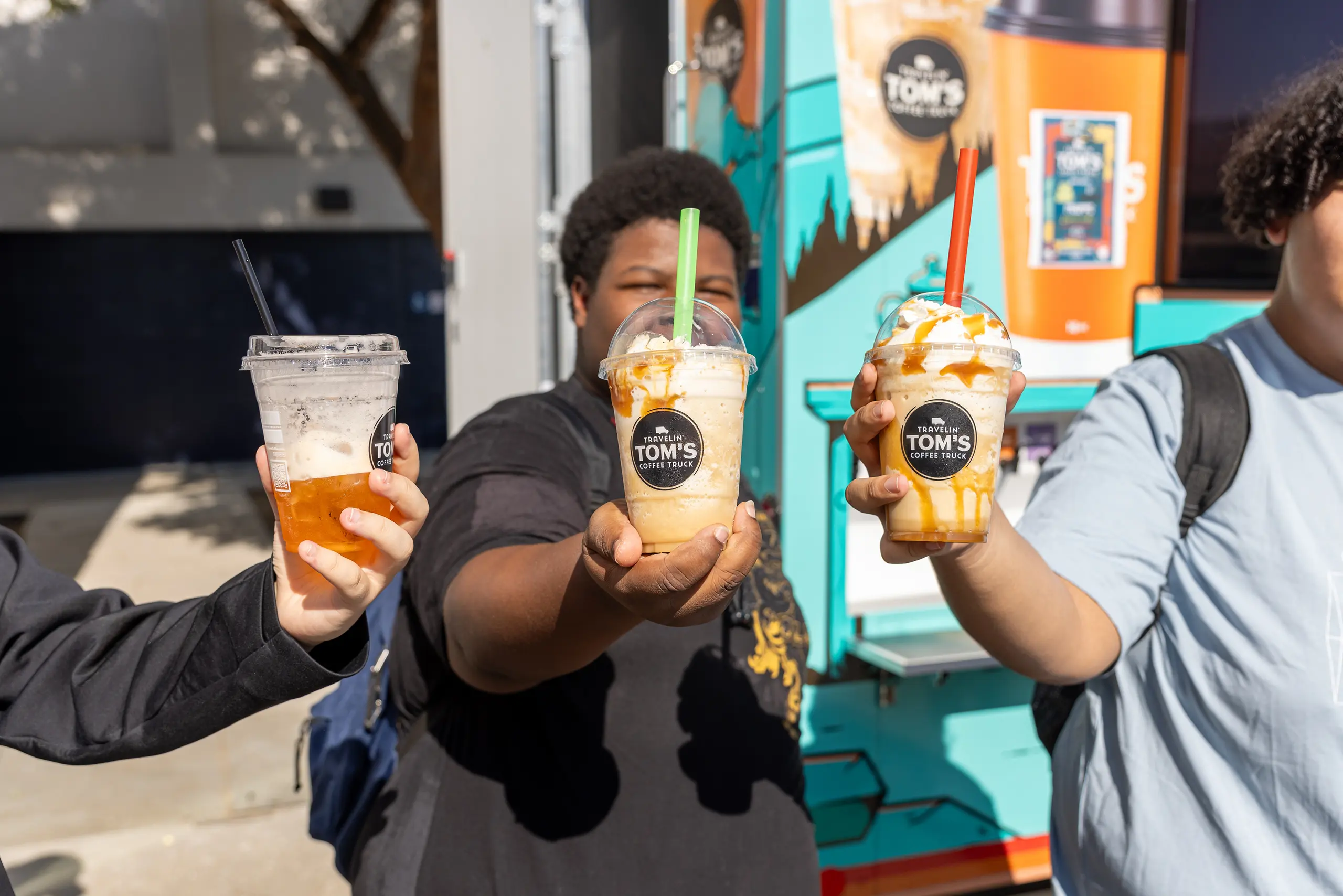 Three people hold up iced beverages in clear cups labeled “Tom’s” in front of a colorful food truck, with two drinks topped with caramel drizzle and whipped cream. The background is outdoors and sunny.
